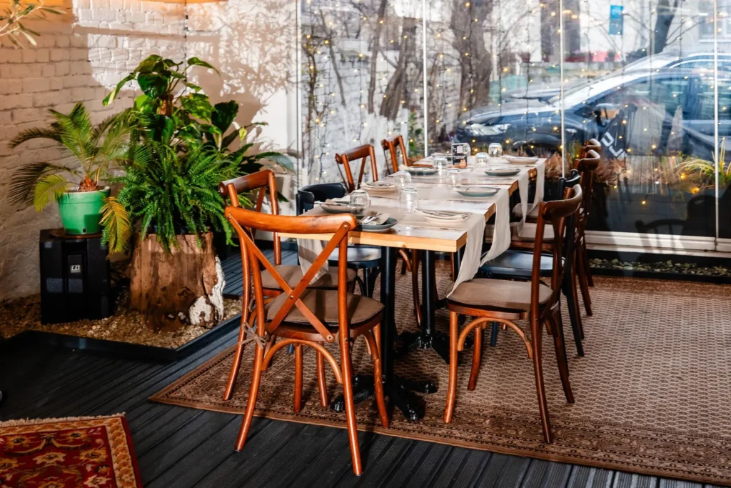 Masă lungă de lemn cu scaune bistro, plante și covor, în sala Brera